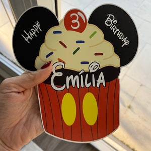 May include: A red and yellow cupcake with a black Mickey Mouse ear design. The cupcake has white frosting with colorful sprinkles and a red cherry on top. The text "Happy Birthday" is written on one ear and "Be Birthday" is written on the other ear. The number "3" is written in red on the cupcake. The name "Emilia" is written in black on the cupcake.