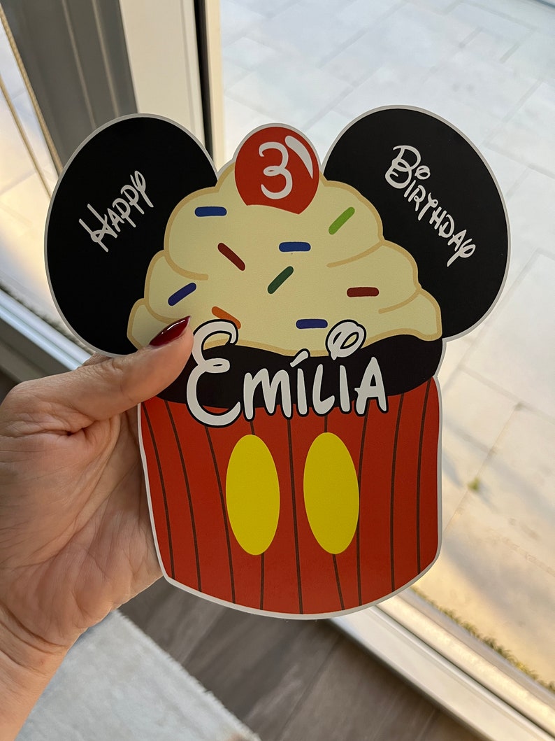 May include: A red and yellow cupcake with a black Mickey Mouse ear design. The cupcake has white frosting with colorful sprinkles and a red cherry on top. The text "Happy Birthday" is written on one ear and "Be Birthday" is written on the other ear. The number "3" is written in red on the cupcake. The name "Emilia" is written in black on the cupcake.