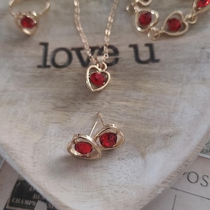May include: A collection of gold-toned jewelry with red heart-shaped gemstones. The set includes a ring, necklace, bracelet, and earrings. The word "love u" is visible on the wooden surface. The jewelry is arranged on a heart-shaped wooden tray.