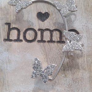 May include: A silver ear cuff featuring five butterfly designs encrusted with sparkling crystals. The ear cuff is set against a wooden background with the word "hom" and a heart symbol.