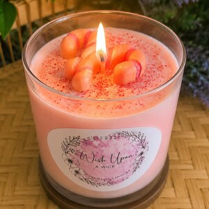 May include: A lit pink candle in a clear glass jar, decorated with candy cane shapes. The candle has a label that reads "Wish Upon A Wick". The candle sits on a brown base.