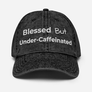 Op de afbeelding: Een zwarte baseballpet met een acid-wash effect en de wit geborduurde tekst Blessed But Under-Caffeinated op de voorpanelen. De pet heeft een gebogen klep, een gestructureerde kroon met gestikte oogjes en een klassieke constructie met zes panelen.