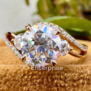 May include: A rose gold engagement ring with a large, round, clear central stone. The ring features a split shank design with smaller accent stones and is set on a brown velvet surface. The text "JD Enterprise" is visible.