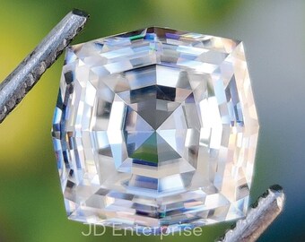May include: A clear, square-shaped gemstone with a faceted design. The gemstone is held by a pair of tweezers.