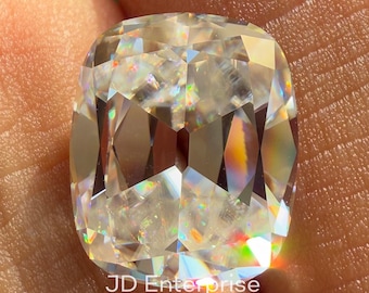 May include: A large, cushion-cut gemstone with a brilliant, multifaceted design. The gem is held between fingers, showcasing its clarity and rainbow-like light refraction. The text "JD Enterprise" is visible on the gem.