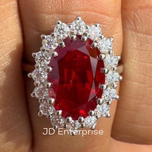 May include: A ring featuring a large, oval-cut, deep red gemstone at the center, surrounded by a halo of small, round, clear, sparkling stones set in a metallic band. The text JD Enterprise is visible below the ring and in the bottom corner.