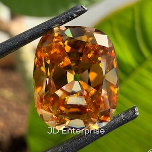 May include: A large, cushion-cut, golden-orange gemstone is held by black tweezers against a backdrop of green leaves. The gem has many facets, reflecting light. The text "JD Enterprise" is visible on the gem.