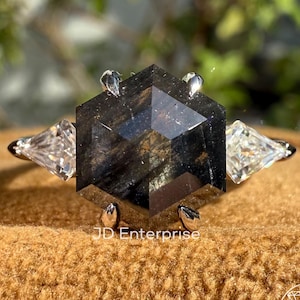 May include: A silver ring featuring a large, hexagonal, dark gray gemstone with a unique pattern. Two smaller, clear, triangular gemstones flank the central stone. The ring is set on a brown surface, with the text "JD Enterprise" visible.