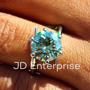 Puede incluir: Un anillo de plata con una gran piedra preciosa redonda de color verde claro. El anillo está engastado con pequeños diamantes alrededor de la piedra preciosa. El anillo está en el dedo de una persona.