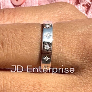 Puede incluir: Un anillo de plata con una banda de pequeñas estrellas incrustadas con pequeñas piedras transparentes. El anillo está en el dedo de una persona.  JD Enterprise.