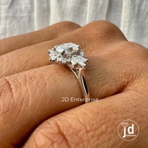 May include: A silver engagement ring with a large oval-shaped center stone surrounded by smaller round stones and marquise-shaped stones. The ring is on a finger.