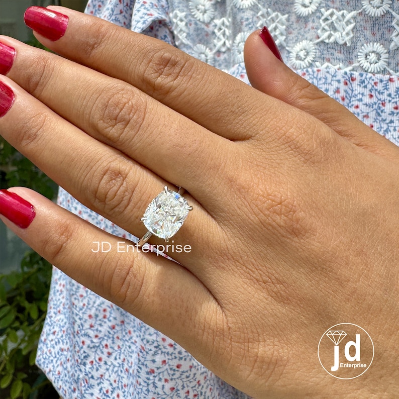 May include: A silver ring with a large, cushion-cut diamond. The ring is on a person's finger.