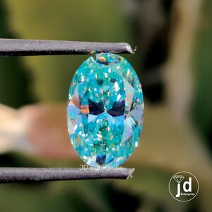 May include: A teal oval-cut gemstone held by tweezers. The gem's facets reflect light, creating a sparkling effect. The image includes the text "JD Enterprise" and a logo.