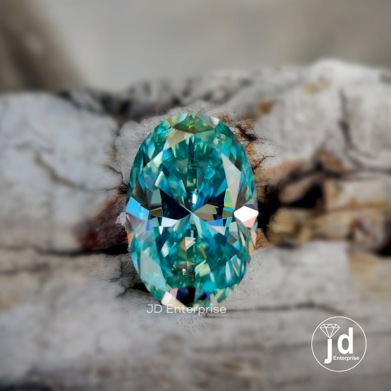 May include: A teal-coloured, oval-cut gemstone with multiple facets. The gem is set against a neutral, textured background. The image includes the text "JD Enterprise" and a logo.