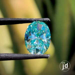 May include: A teal-coloured, oval-cut gemstone held by tweezers. The gem's many facets reflect light, creating a sparkling effect. The text "JD Enterprise" is visible on the tweezers.