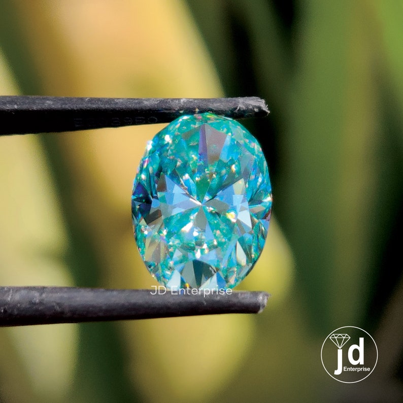 May include: A teal-coloured, oval-cut gemstone held by tweezers. The gem's many facets reflect light, creating a sparkling effect. The text "JD Enterprise" is visible on the tweezers.