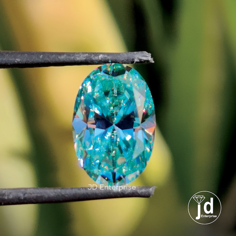May include: A teal-coloured, oval-cut gemstone held by black tweezers. The gem's facets reflect light, creating a sparkling effect. The background is a blurred green and yellow. The text "JD Enterprise" is visible.
