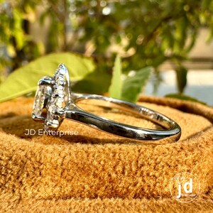 May include: A silver ring with a large, clear, oval-shaped gemstone surrounded by a halo of smaller gemstones. The ring is on a brown velvet box with the text "JD Enterprise" visible.