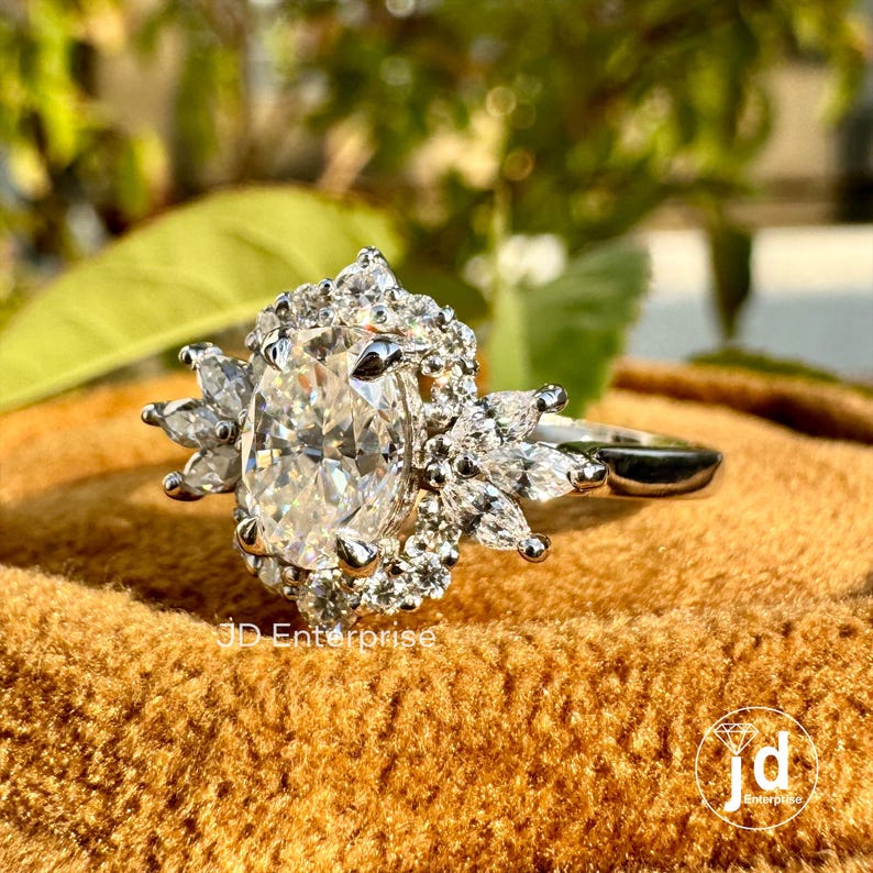 May include: A silver ring with a large oval-shaped clear gemstone surrounded by smaller clear gemstones. The ring is set on a brown velvet surface.
