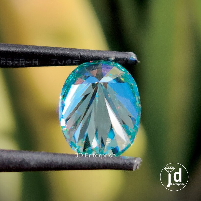 May include: A light blue, oval-cut gemstone with detailed facets is held by black tweezers. The gem displays a range of colours, including blue, green, and purple. The background is a soft, blurred green.
