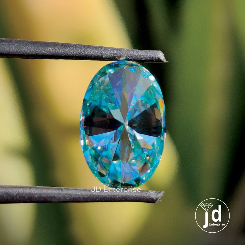 May include: A teal-coloured, oval-cut gemstone held by tweezers. The gem's facets reflect light, creating a sparkling effect. The text "JD Enterprise" is visible at the bottom of the image.