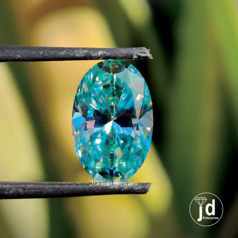 May include: A teal-coloured, oval-cut gemstone held by black tweezers. The gem's many facets reflect light, creating a sparkling effect. The background is a blurred green and yellow.