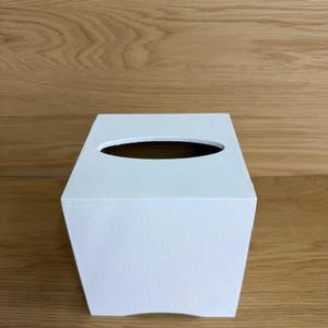 May include: A white, rectangular tissue box with a curved top and an oval opening. The box is painted white and has a slightly textured surface. The tissue box is made of wood and is designed to hold a standard box of tissues.