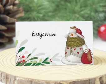 Puede incluir: Una tarjeta de lugar de Navidad imprimible con una ilustración de muñeco de nieve con un sombrero y una bufanda rojos, acebo verde y el nombre "Benjamin".