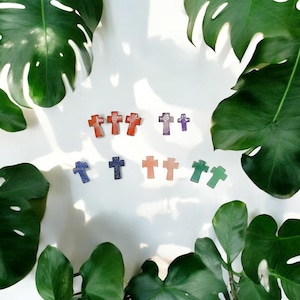 May include: A collection of small, colorful cross-shaped charms made of stone. The charms are arranged in rows on a white background. The charms are in shades of red, orange, purple, blue, green, and pink.