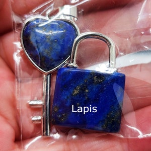 Puede incluir: Un colgante de llave en forma de corazón y un candado cuadrado de lapislázuli azul oscuro. La llave tiene un marco y detalles de color plateado. El candado tiene un asa de color plateado. La palabra "Lapis" está impresa en el candado.