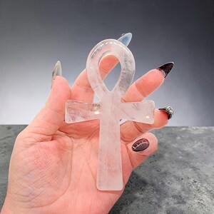 May include: A carved ankh symbol made of translucent, pale pink rose quartz. The object features a smooth, polished surface with natural internal inclusions and a classic cross shape with a looped top, held upright to display its clear, crystalline structure.