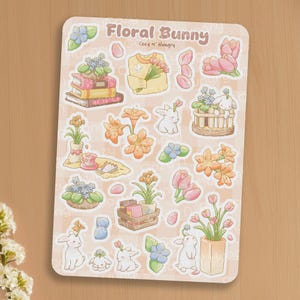Peut inclure: Une planche d'autocollants sur le thème du lapin floral. Le design comprend des illustrations de lapins, de fleurs, de livres et d'un pull. Le texte "Floral Bunny" et "Cozy N' Hungry" est en haut. La palette de couleurs est douce, avec des roses, des jaunes et des verts pastel.