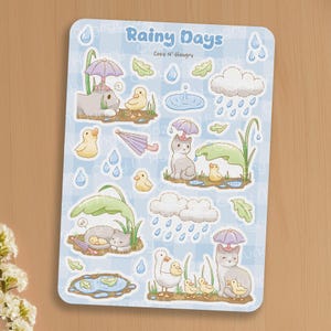 May include: A sticker sheet with illustrations of cats and ducks in rainy scenes. The title "Rainy Days" is in blue script, with "Cozy N' Hungry" below. The illustrations include umbrellas, rain clouds, and puddles, set against a light blue checkered background.