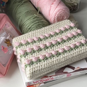 Könnte beinhalten: Eine handgefertigte Häkeltasche in Creme mit rosa und grünen floralen Akzenten liegt auf einem Manga-Buch. Zwei Wollknäuel, eines rosa und eines grün, sind sichtbar, zusammen mit einem rosa Beutel und einer Hello Kitty-Figur.