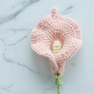 Może przedstawiać: Ręcznie robiony szydełkowy kwiat kalii w delikatnym różu, z kremowym środkiem i zieloną łodygą. Kwiat wykonany jest z teksturowanego ściegu, ukazującego szczegóły przędzy. Tło to biała marmurowa powierzchnia.