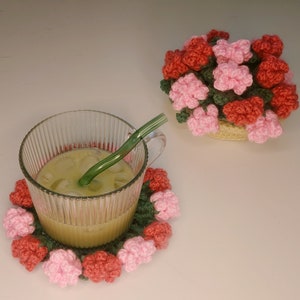 Puede incluir: Una taza de vidrio transparente con una pajita verde descansa sobre un posavasos de ganchillo con detalles de flores rojas, rosas y verdes. También hay una cesta de ganchillo llena de flores rojas y rosas.