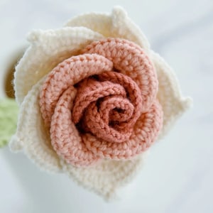 May include: A handmade crochet rose in a white vase. The flower features a layered design with a cream-colored outer layer and a peach-colored center. The flower is a decorative item.