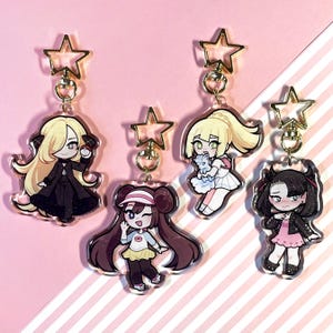 May include: Four colorful anime-style character keychains with gold star-shaped charms. The characters are a girl with blonde hair and a white dress, a girl with brown hair and a pink shirt, a girl with black hair and a black dress, and a girl with black hair and a pink shirt.