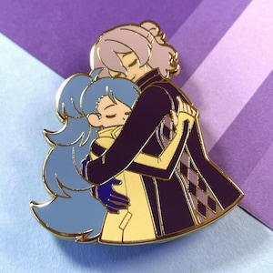 Peut inclure: Épinglette en émail doré représentant deux personnages, l'un avec des cheveux violets et une veste à carreaux violets et blancs, en train d'embrasser un personnage avec des cheveux bleus et une veste jaune.