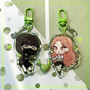 Op de afbeelding: Twee sleutelhangers met cartoonfiguren en groene sleutelringen. De sleutelhanger aan de linkerkant heeft een personage met zwart haar, een zwart masker en een zwart-witte outfit. De sleutelhanger aan de rechterkant heeft een personage met roze haar, een wit shirt en een zwarte rok. Beide personages lachen.