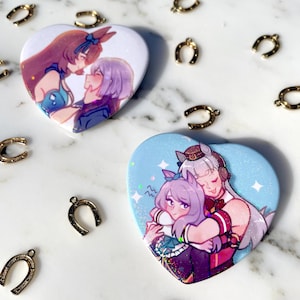 May include: Two heart-shaped buttons with anime-style illustrations of women. One button features two women embracing, set against a light blue background with sparkles. The other button shows two women in a romantic pose. Gold horseshoe charms are scattered around.