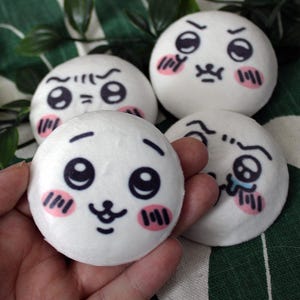 Pode incluir: Quatro brinquedos de pelúcia redondos brancos com rostos de desenhos animados expressivos. Cada um tem olhos, sobrancelhas e boca pretas, com marcas de blush rosa nas bochechas. Um é segurado na mão, e os outros estão dispostos em uma superfície estampada verde e branca.