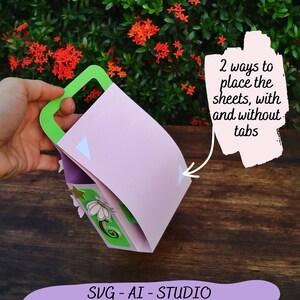 May include: A pink paper craft box with a green handle. The box is folded and has white tabs on the sides. The text "2 ways to place the sheets, with and without tabs" is written on a white background.