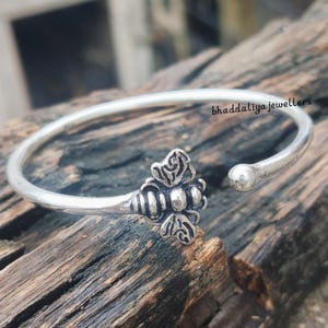 Pode incluir: Uma pulseira de punho prateada com um design de abelha. A pulseira apresenta um design aberto com uma pequena bola numa extremidade. A abelha tem asas e corpo detalhados. A pulseira é exibida em um pedaço de madeira envelhecida.