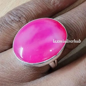 May include: A large, oval-shaped ring with a vibrant pink gemstone set in a silver band. The gemstone has a smooth, polished surface. The text "laxmisilverhub" is visible on the ring.