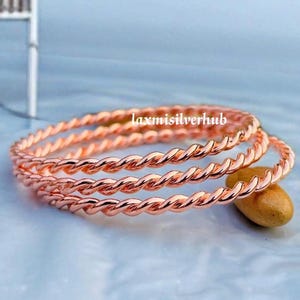 Puede incluir: Un conjunto de tres brazaletes de metal trenzado en tono oro rosa con una textura continua similar a una cuerda. Las pulseras están dispuestas en una pila y el texto laxmisilverhub es visible en el centro de la imagen sobre las piezas.