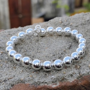 May include: A silver bracelet featuring a series of large, spherical beads. The polished metal beads reflect light, creating a bright appearance. The bracelet is displayed on a textured, grey surface. The beads are evenly spaced.