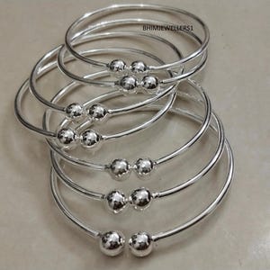 Op de afbeelding: Verschillende zilveren armbanden met een eenvoudig ontwerp. Elke armband heeft een gladde, afgeronde band en is versierd met drie kleine, gepolijste zilveren kralen. De armbanden zijn gestapeld, waardoor hun reflecterende, glanzende oppervlak zichtbaar is.