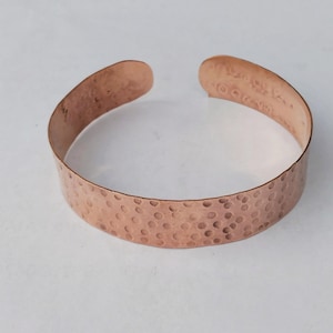Puede incluir: Una pulsera de puño de cobre con textura martillada. La pulsera abierta tiene un color marrón rojizo cálido y una serie de pequeñas hendiduras espaciadas uniformemente en su superficie. La pulsera está diseñada para usarse en la muñeca.