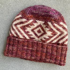 Könnte beinhalten: Eine gestrickte Mütze mit einem geometrischen Muster in Braun-, Creme- und Rosatönen. Die Mütze hat einen umgeschlagenen Rand.